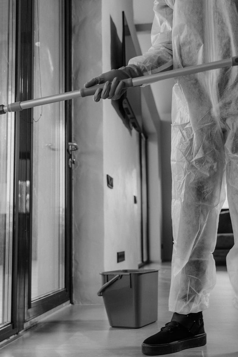 A person in a protective suit cleans glass doors indoors, emphasizing hygiene.